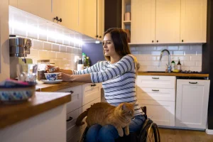 Disabled woman in wheelchair in kitchen making a cup of tea with cat sitting on lap
